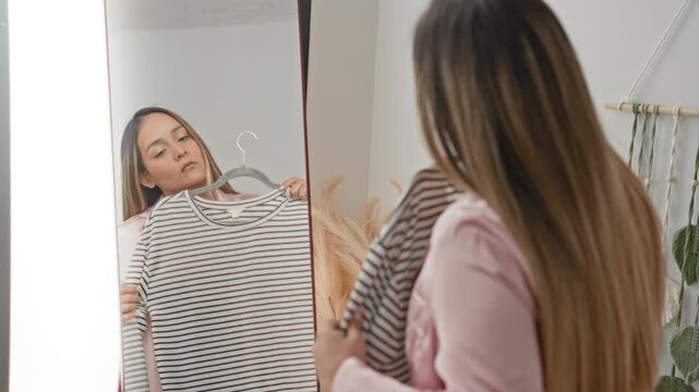 Woman wearing pink pajamas holds striped shirt on hanger before fulllength mirror in studio; contemplation decision.