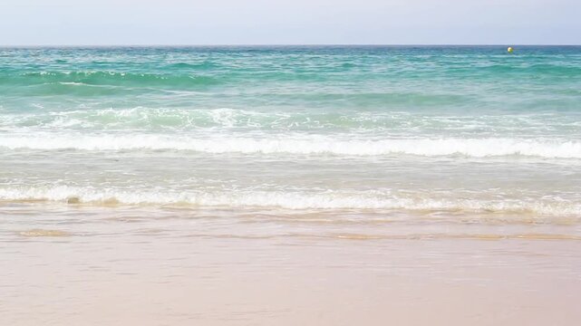 Rear view of a person running into shallow ocean waves from a sandy beach with turquoise water and sea spray under a clear horizon.