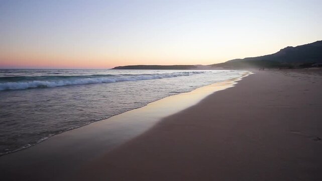 Ocean waves rolling onto a sandy beach at sunset with a curving coastline and distant headland along the Spanish coast under a clear evening sky.