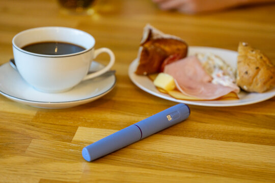 A blue injection pen for diabetes or weight loss treatment placed on a wooden table next to a cup of black coffee and a breakfast plate, symbolizing daily medication routine and healthy lifestyle.