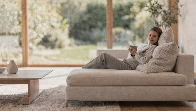 Young woman relaxing on a comfortable modern sofa in a bright, minimalist living room with garden views