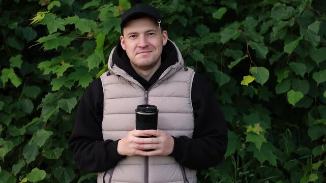 Man standing in park holding coffee cup in hand. Caucasian young male wearing cap and vest looking away. Relaxing in nature outdoors. Casual lifestyle and coffee break concept.