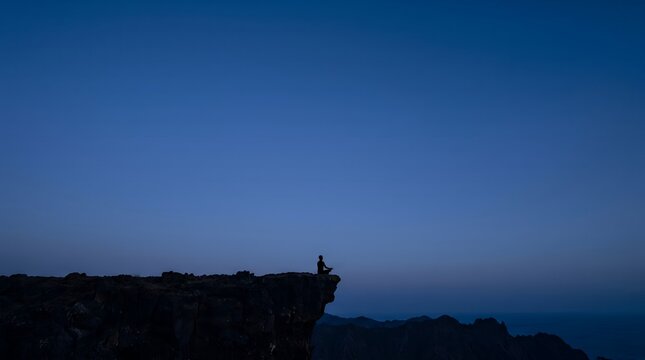 A person stands on the edge of a cliff overlooking a vast landscape at dusk.
