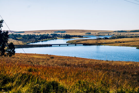 Tranquil river landscape with a concrete bridge crossing a wide blue river surrounded by golden winter grass and rolling hills under a clear blue sky, eMalahleni, South Africa.