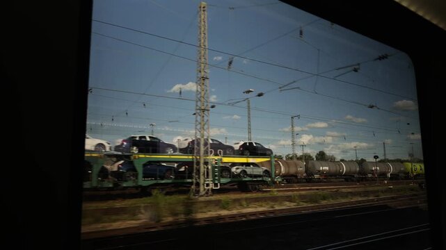 Passenger perspective through a railway window capturing a cargo train transporting manufactured vehicles near a logistics hub. Highlights Bavarian industrial and rail infrastructure.