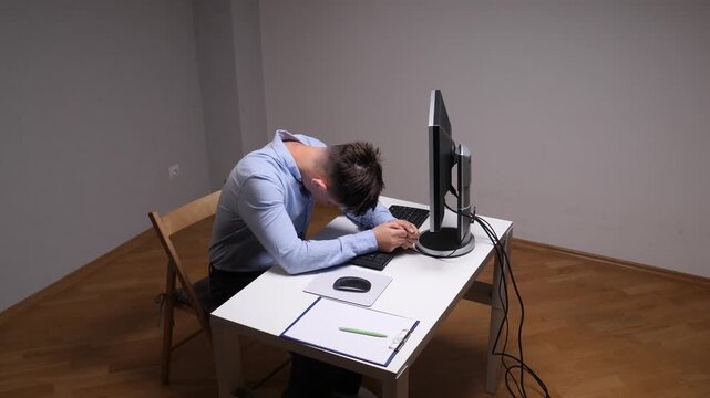 Young man sleeping head down at his office desk late at night, exhausted from long hours and overwork, showing burnout, stress and fatigue while working on a computer