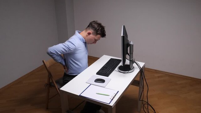 Young man experiencing discomfort and holding his lower back while working at a computer in an office, illustrating issues like poor posture, sedentary lifestyle, and prolonged screen time