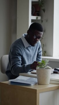 Vertical dolly away shot of young adult African American man wearing glasses, writing in notebook and working on computer in office. Footage suitable for corporate training or tech recruitment videos