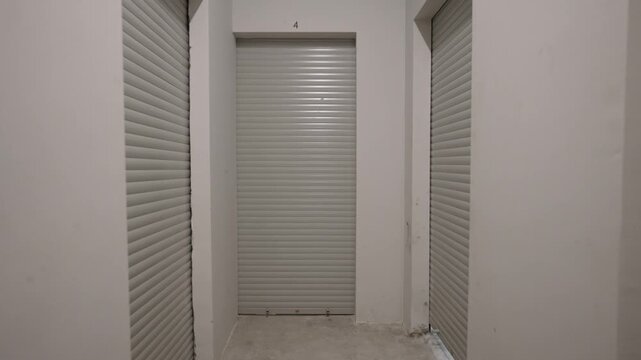 Video walkthrough of a clean storage facility hallway showing multiple closed roller shutter doors in numbered units with white walls and concrete flooring