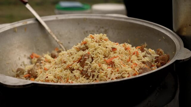 Cook stirring pilaf in a large iron cauldron over an open fire. Traditional rice dish with meat and carrot being prepared in a heavy pot outdoors for meal.