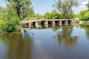 Obraz premium Old concrete low bridge crossing a calm river surrounded by green trees on a sunny summer day