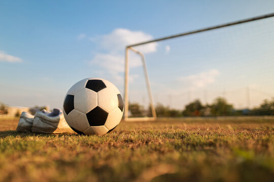 Football training and active lifestyle concept. Close up of a classic soccer ball and football boots on green grass field with blurred goal net in the background at sunset.