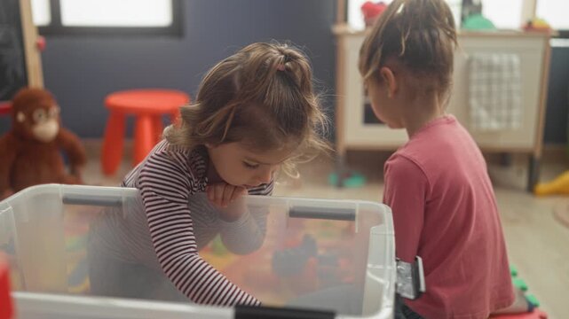 Girl reaches into a clear plastic toy bin with hand and picks up blocks in a colorful playroom; quiet curiosity.