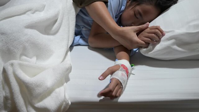 Caring mother holding hand and touching forehead of sick asian little girl patient resting on bed in hospital room. Healthcare, family love, medical recovery and pediatric support concept.