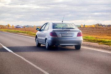 Naklejka premium Silver sedan car driving away on rural asphalt road through countryside under cloudy sky, fields with dry grass and autumn trees along road