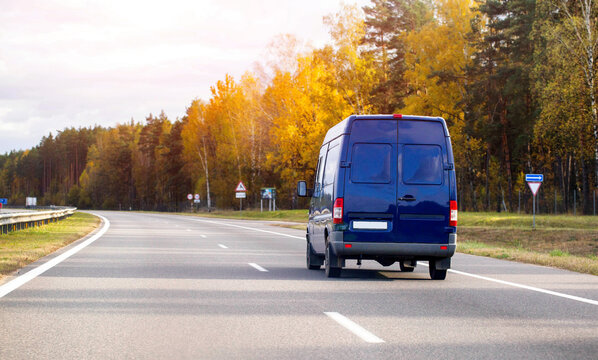 Blue delivery van traveling on asphalt highway through autumn forest, vehicle moving on open road surrounded by colorful fall trees under bright sky, daytime transportation