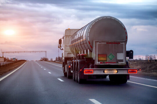 White tanker truck driving on asphalt highway at twilight, transporting hazardous liquid, rear view of vehicle on road under cloudy sky, evening sunset. Copy space for text
