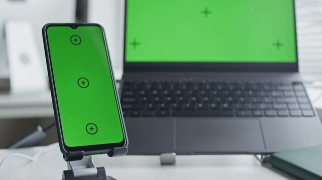 Smartphone on stand and laptop computer displaying green screens with tracking markers sitting on office desk. Mockup template ideal for showcasing mobile application or software design