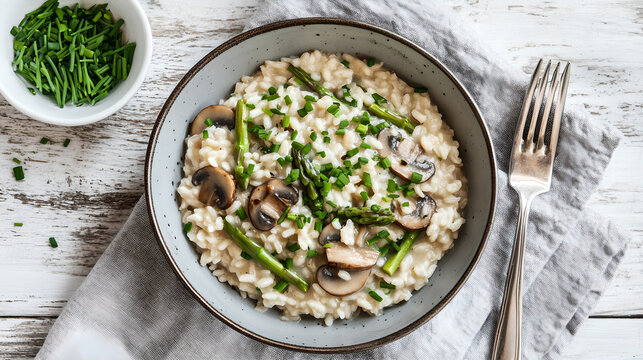 Top view of a bowl of creamy mushroom and asparagus risotto garnished with fresh chives on a rustic white wooden table