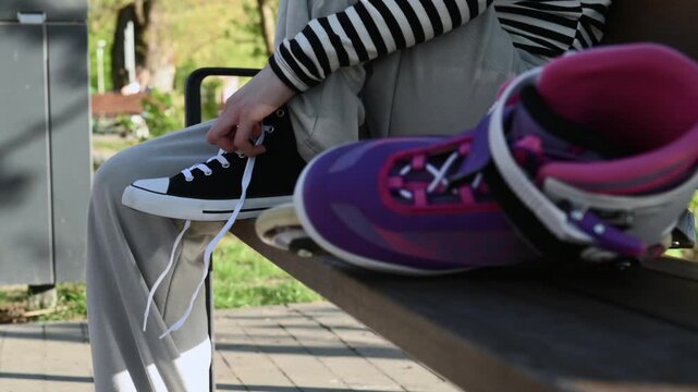Teen girl sits on a park bench tying black shoelaces A purple roller skate waits nearby in the sunny outdoor park