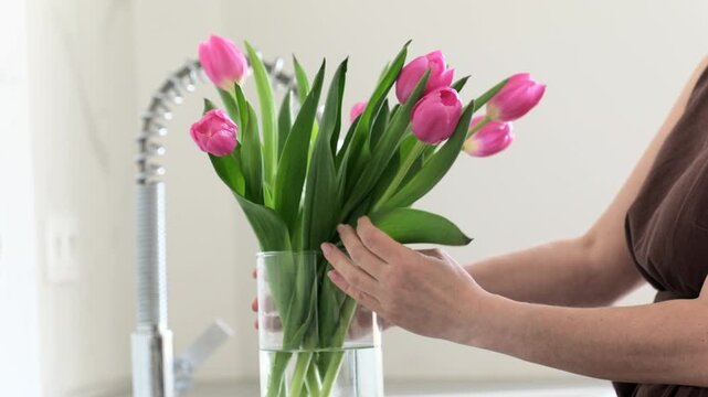 Woman carefully arranges fresh pink tulips in clear glass vase on bright kitchen counter.