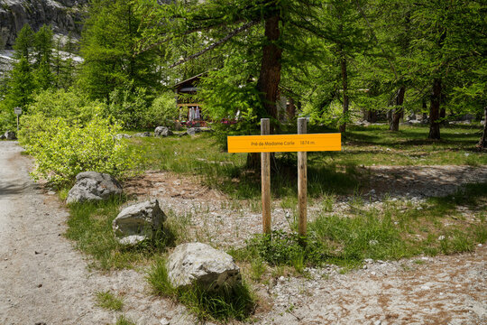 A yellow trail sign reading &ldquo;Pr&eacute; de Madame Carle 1874 m&rdquo; in a summer high-mountain landscape. In the background stand numerous larch trees, among which is a mountain hut with a terrace.