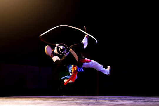 Dynamic Korean pungmul performer spinning sangmo ribbon while jumping with a small drum on a dark stage.