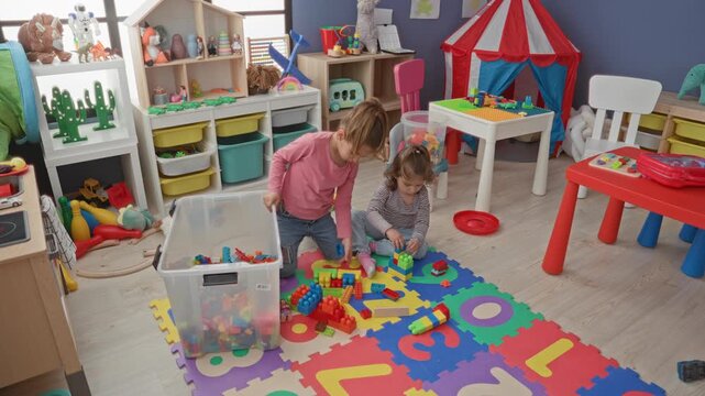 Two girls stacking colorful plastic blocks while sitting on foam alphabet mat inside building; playful friendship.