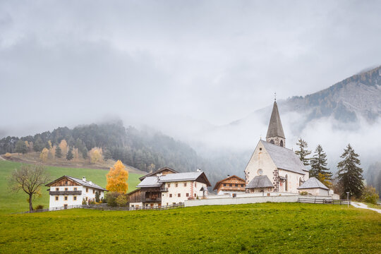 Church Chiesa di Santa Maddalena stands beneath low clouds and misty mountain slopes in Val di Funes, South Tyrol. Atmospheric autumn landscape in the Dolomites