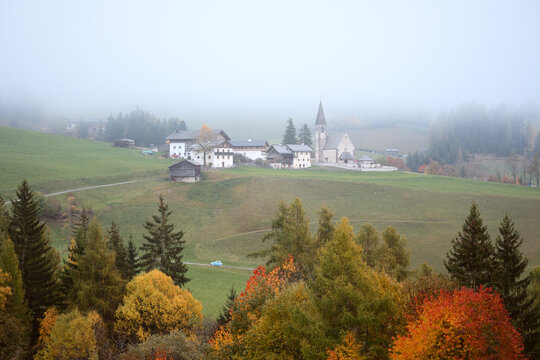 Chiesa di Santa Maddalena surrounded by autumn forests and low clouds in Val di Funes. Scenic rural landscape in the Dolomites of northern Italy