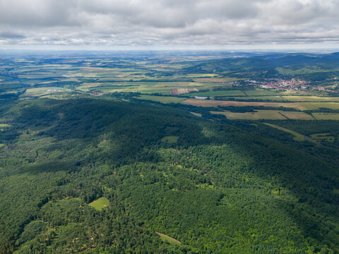 Aerial view of green forests of Bohunicky Rohac in the Stiavnicke Vrchy mountains with agricultural fields and a village under a cloudy sky in Bohunice, Nitra Region, Slovakia.