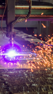 Automated industrial machine cutting thick metal plate with high-power plasma arc and brilliant purple light, producing vibrant orange sparks flying in manufacturing workshop vertical photo