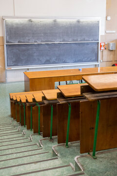 Empty university lecture hall with rows of wooden desks and seats arranged on tiered steps, facing a large double blackboard on the wall in an old traditional classroom setting for higher education.