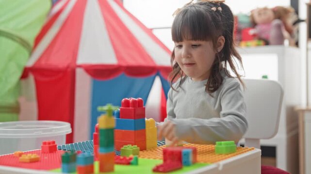 Little girl stacking colorful plastic blocks on a play table in a bright indoor playroom with tent and toys; concentration.