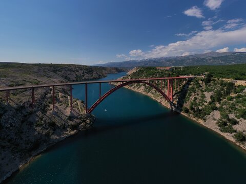 Aerial view of Maslenica bridge, a red arch structure spanning the deep blue water of the Maslenica Strait, surrounded by rugged rocky cliffs and green hills in Jasenice, Croatia.