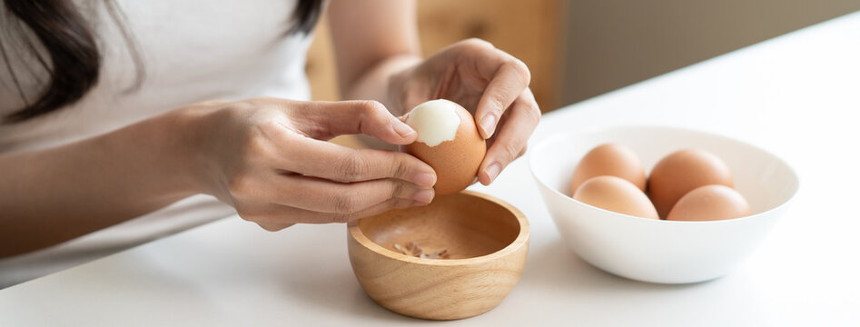 Close up of woman peeling boiled egg at white table, female hands cracking brown egg shell for healthy breakfast meal, high protein food and nutrition concept
