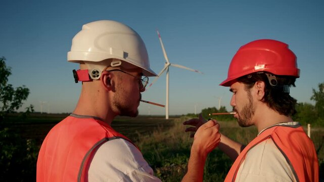 Two industrial workers in protective helmets and high visibility vests pause work at renewable energy site. Cigarettes in hands and wind turbines in background show casual break during project