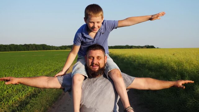 Little boy sitting on shoulders of his daddy and playing raised hands as airplane at sunset. Happy father and his son having fun at countryside. Dad with small kid spending time together outdoor