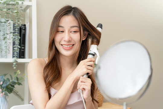 Beautiful young Asian woman curling her hair with curling iron looking in mirror at home, smiling female influencer getting ready creating hairstyle tutorial for lifestyle blog