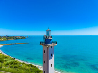 Lighthouse stands tall by the shoreline with blue water and clear sky in a coastal area during daytime © DSMT