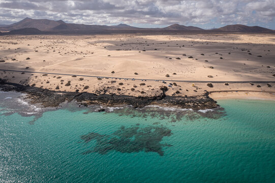 Aerial view of the vast sand dunes of Parque Natural de Corralejo meeting the turquoise ocean and volcanic rocky shore under a cloudy sky in Corralejo, Canary Islands, Spain.