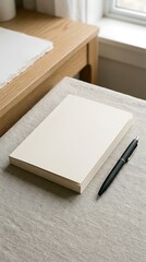 Fototapeta premium A blank notebook rests on a neutral surface next to a pen, with a wooden table and a window in the background