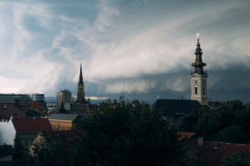Obraz premium City skyline with church steeple and cloudy sky over rooftops, urban architecture under calm morning light glow.