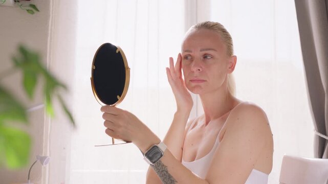 Middle aged woman examining reflection in mirror and checking facial skin, focusing on wrinkles and beauty self care routine at home.Beauty routine , skincare , self care concept