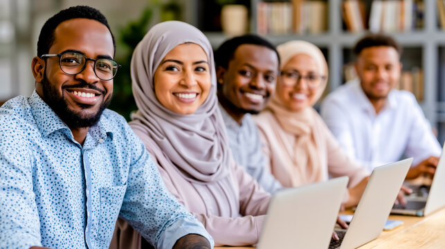 A group of four people two men and two women are smiling and sitting at a table with laptops in front of a bookshelf