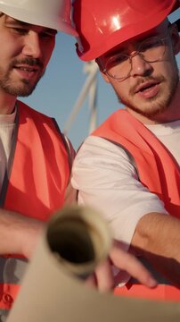 Two European construction workers carefully review and discuss wind turbine blueprints during installation process at wind farm site. Engineers in safety vests and hard hats examine rolled technical