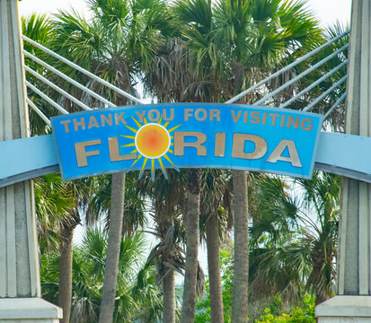 A colorful arch sign reading 'Thank You For Visiting Florida' framed by tall palm trees and bright sun graphics, evoking travel, coastal tourism and a warm tropical vacation atmosphere.