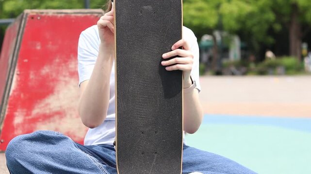 Girl sitting in skate park, holding skateboard in front of her. Front view of deck with black griptape. Teenager resting after skateboarding. Concept of active recreation, street culture and youth lei