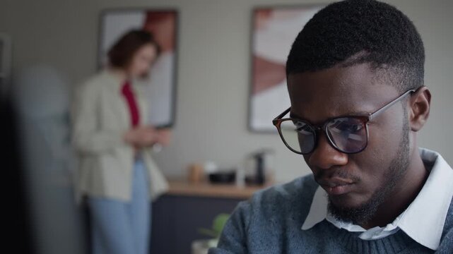 Rack focus shot of Black young adult man wearing glasses focusing on computer screen while Caucasian young adult woman pouring coffee in background. For corporate culture and IT team promotions