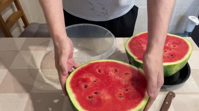 White person arranging watermelon halves on table, placing bowl and plate, flipping half into bowl to drain, revealing juicy red flesh and seeds, knife and patterned tablecloth visible, relaxed